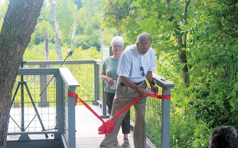 Press photo/Thomas Sherrill - Macon County Board of Commissioners Chair Gary Shields unties the ribbon for the Barbara McRae Memorial Viewing Platform with Friends of the Greenway member Rita St. Clair during the June 23 dedication event.